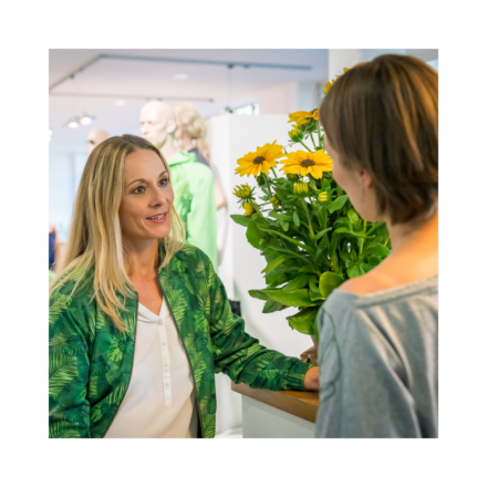 Holfelder Mitarbeiterin in Corporate Wear berät Kundin; Blumenstrauß im Hintergrund